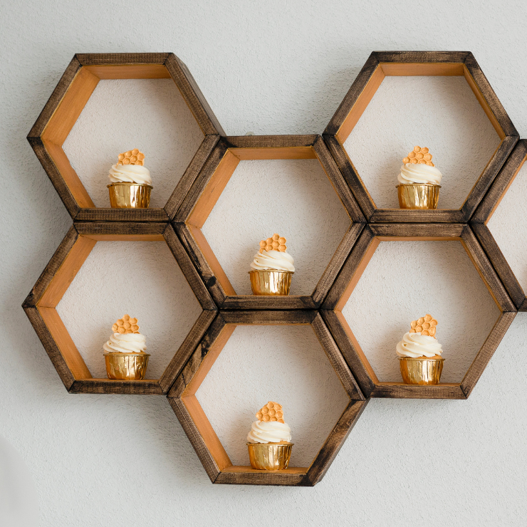Bride to bee bridal shower cupcakes in gold foil patty cases with white cream swirls and honeycomb wafers on a wooden honeycomb shaped shelf.